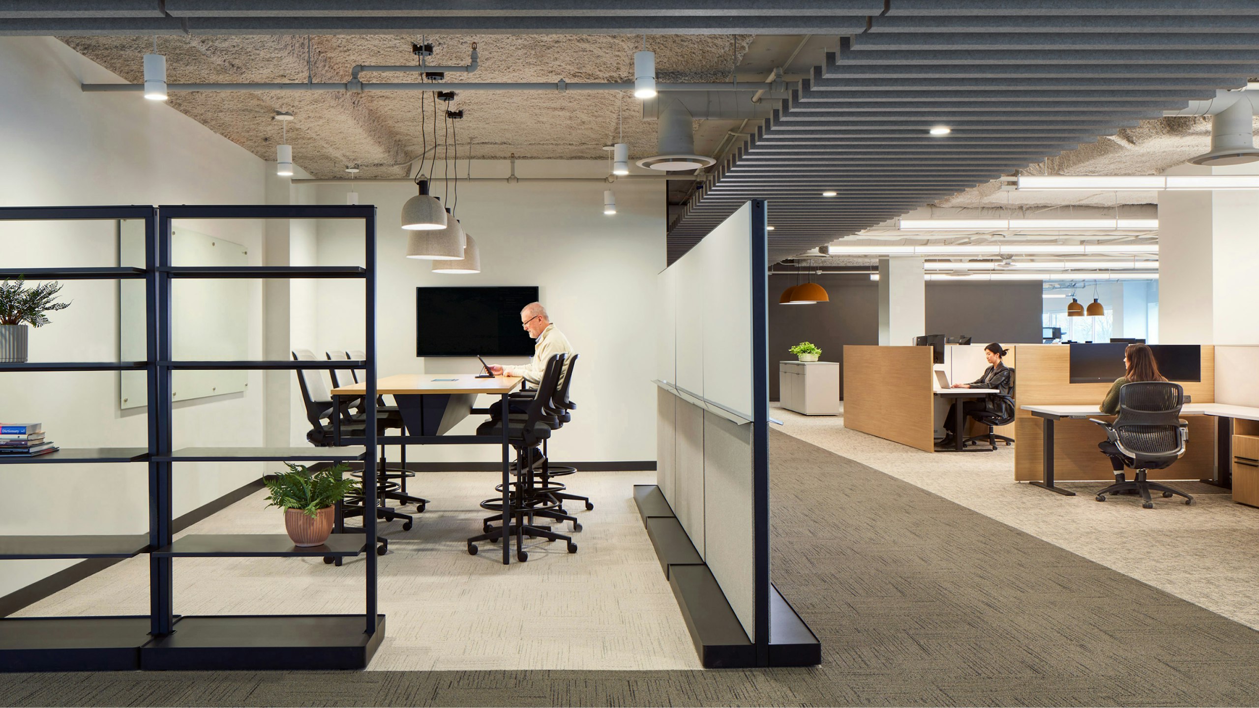 A person sits in a small conference room furnished with counter height Knoll Ollo stools and a table. Two others sit at private workstations in an open office setting.
